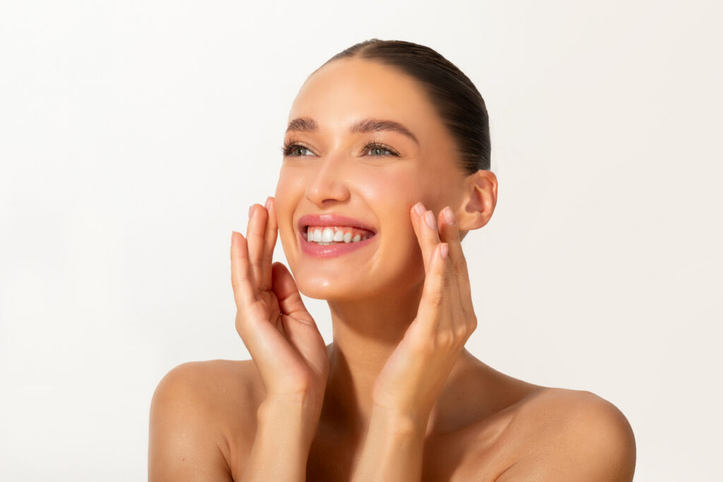 Face skincare delight. Beautiful young lady touching smooth and soft skin and smiling, posing on light background, free space