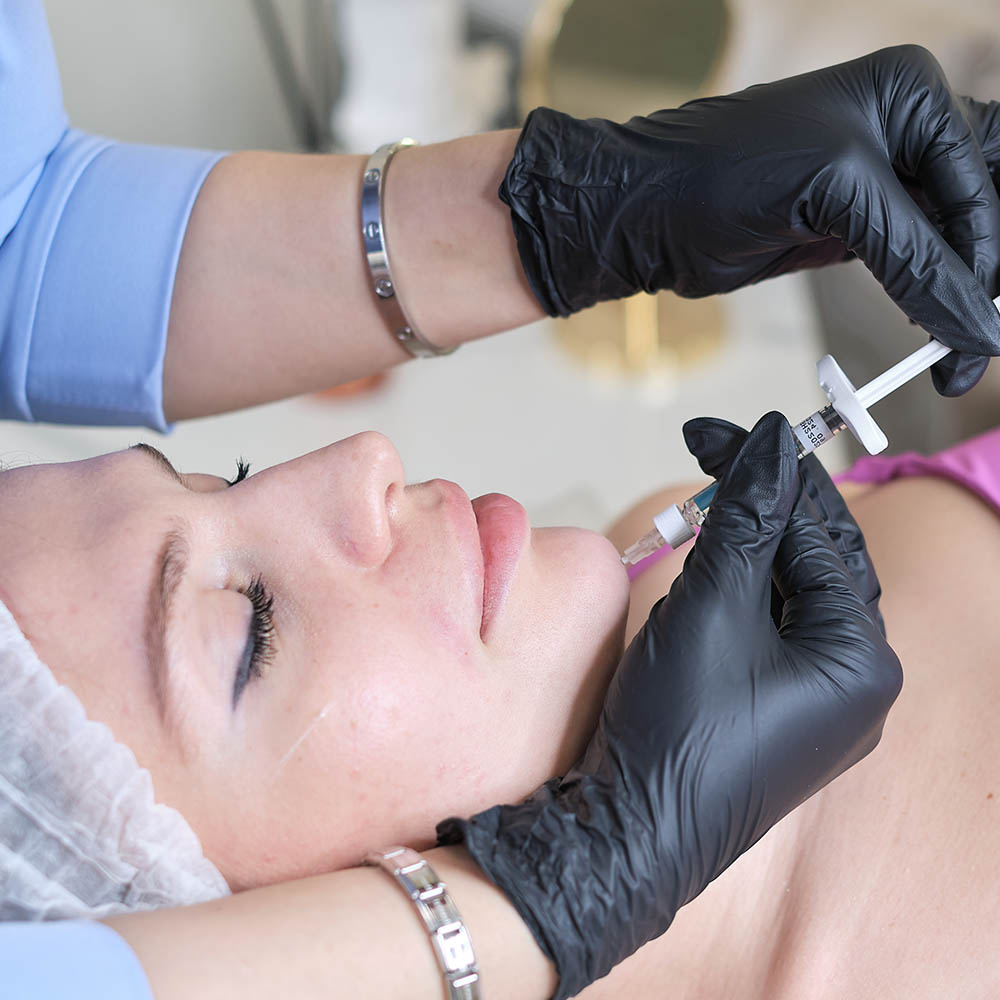 Clinician in black gloves administers a facial injection to a patient lying down during a cosmetic treatment.