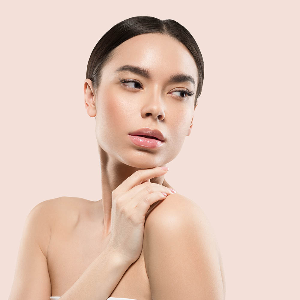 Close-up of a woman with smooth skin gazing to the side, touching her shoulder against a pale pink background.